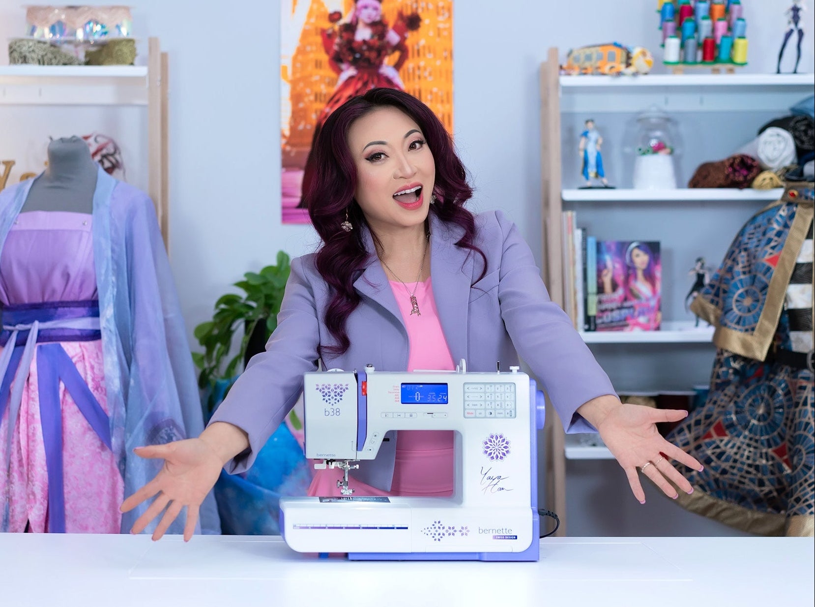 A woman with dark hair wearing a lavender blazer and pink top sits behind a white and lavender sewing machine on a table, with shelves of sewing supplies and a mannequin in a purple dress in the background.